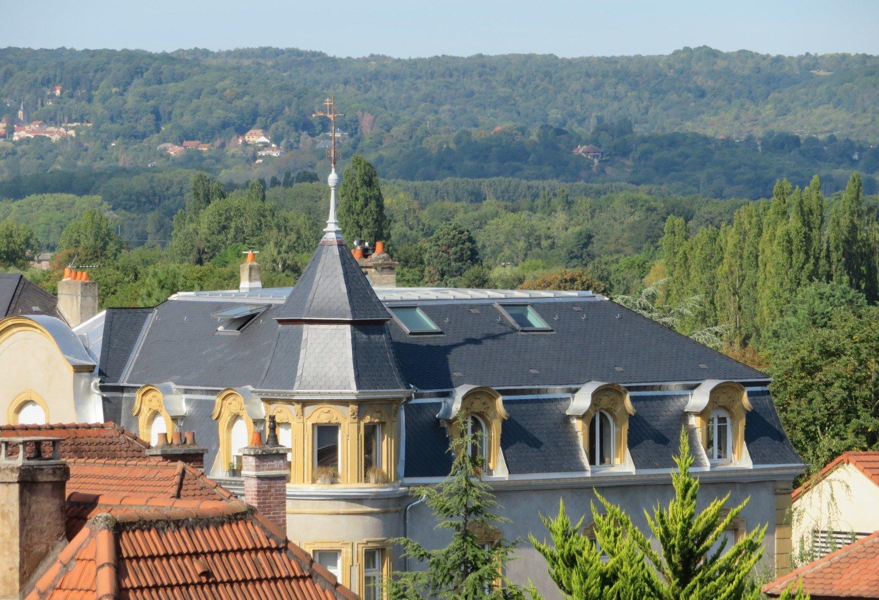 Grande toiture en ardoise avec tourelle surmontée d'un épi, fenêtres de toit et lucarnes sculptées, devant les collines