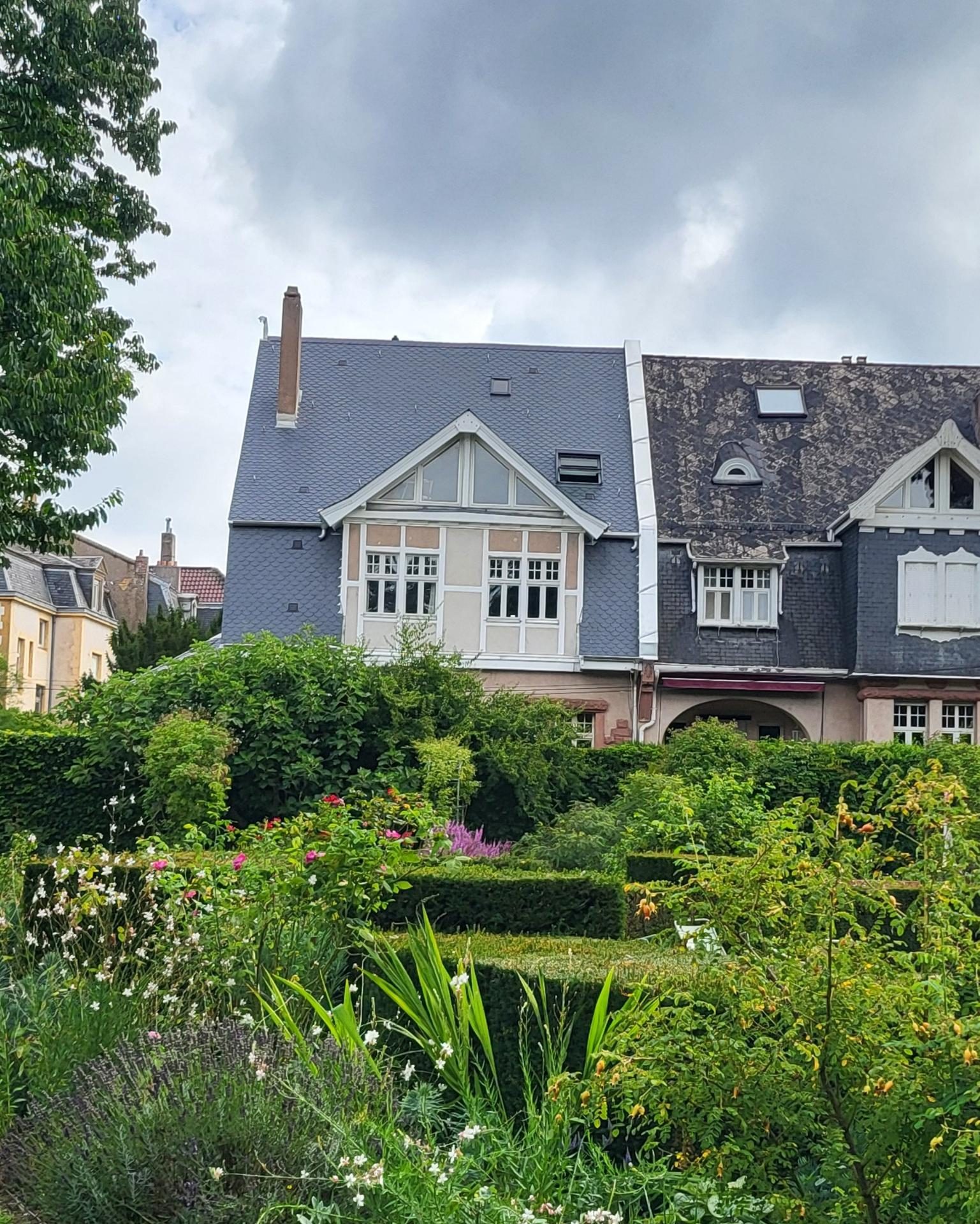 Maison au toit d'ardoise en écailles neuves, à côté de la toiture voisine ancienne, derrière un jardin fleuri